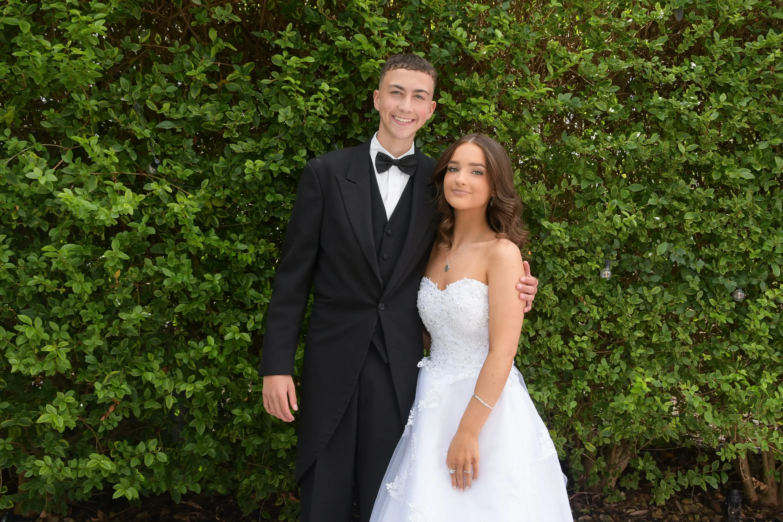 A young couple in formal debutante attire posing together in front of a green hedge.
