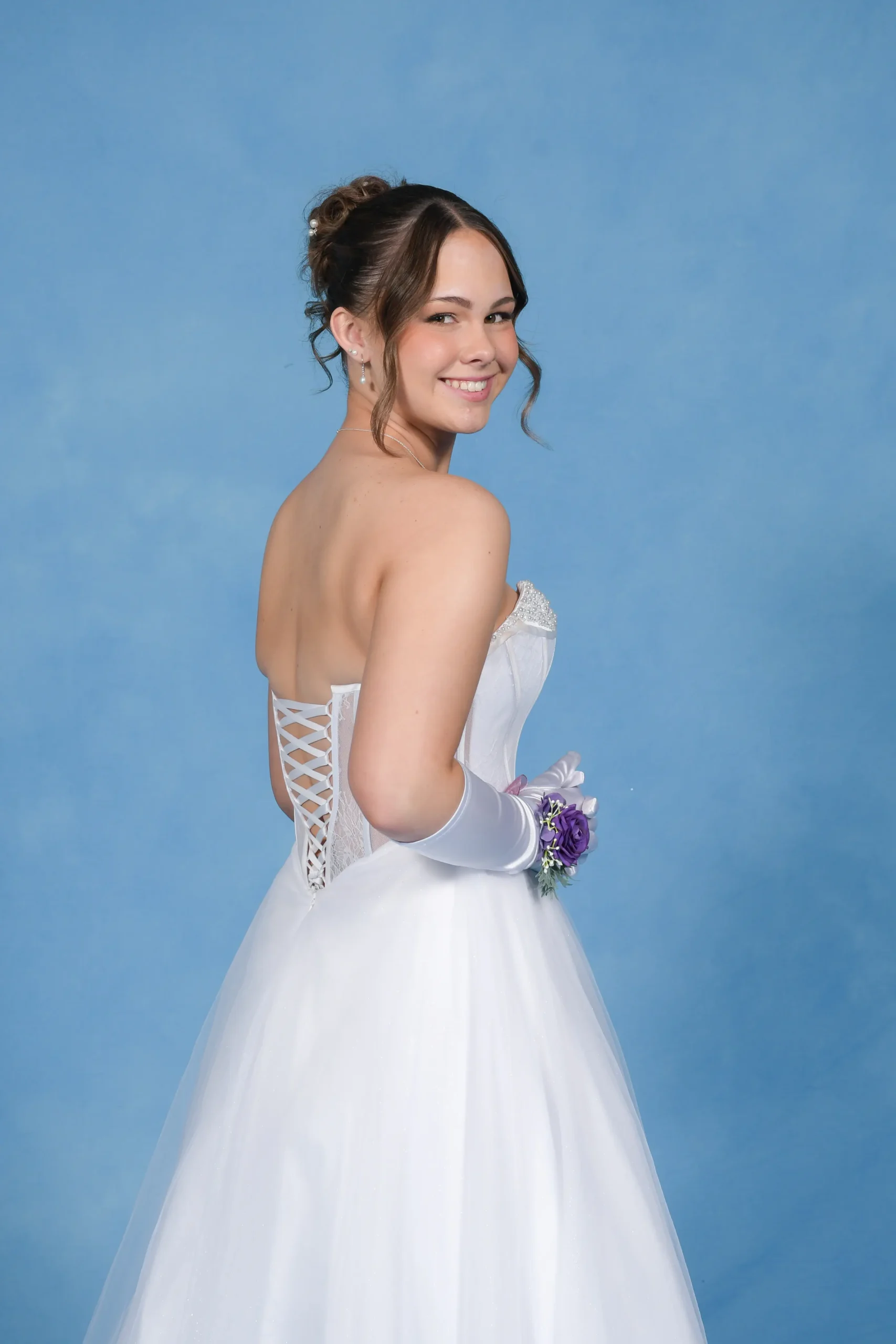 A smiling debutante in a white gown and gloves looking back over her shoulder against a blue background.