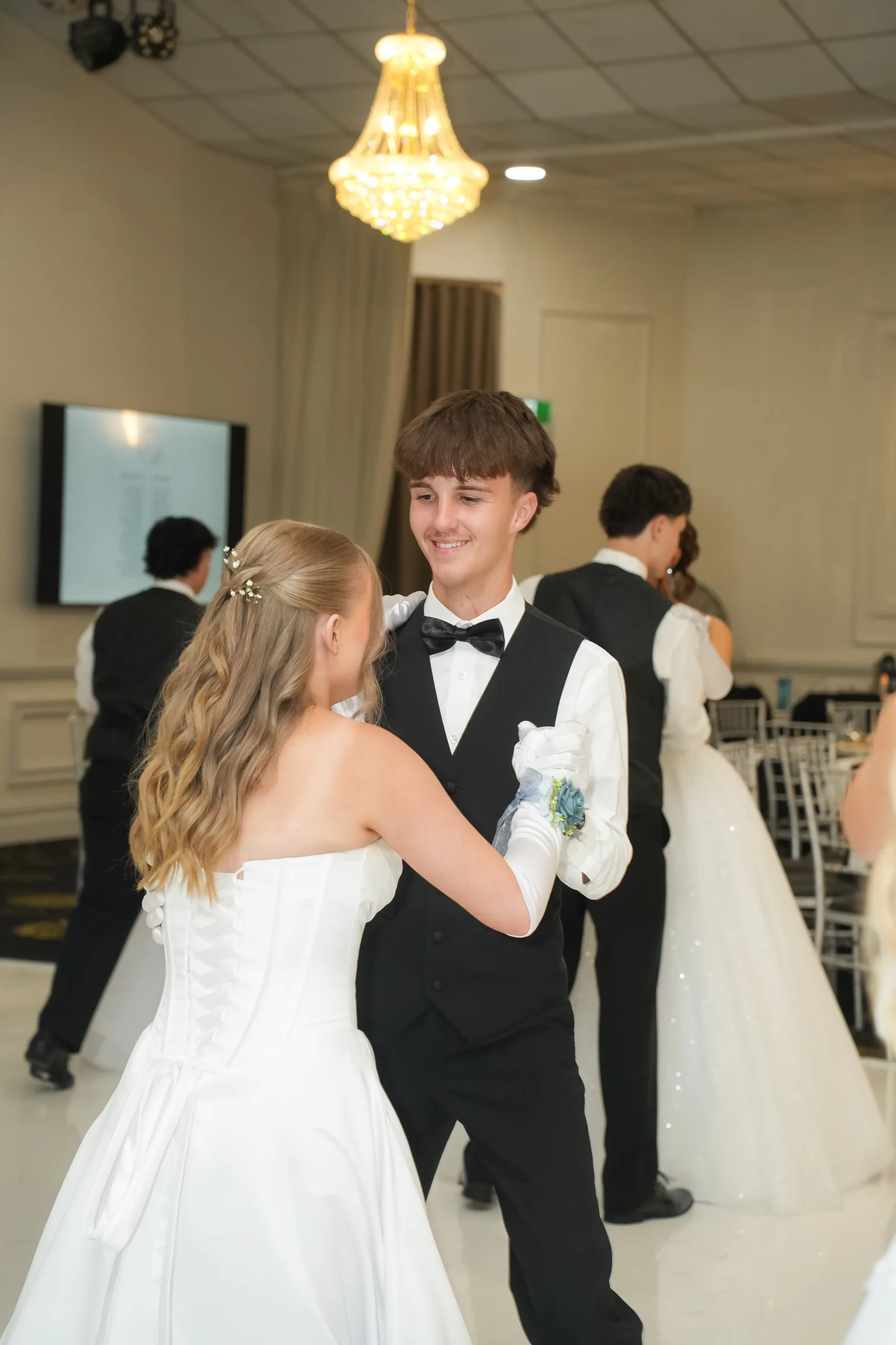A smiling young man in a black vest and bow tie dancing with a debutante in a white gown under a ballroom chandelier.