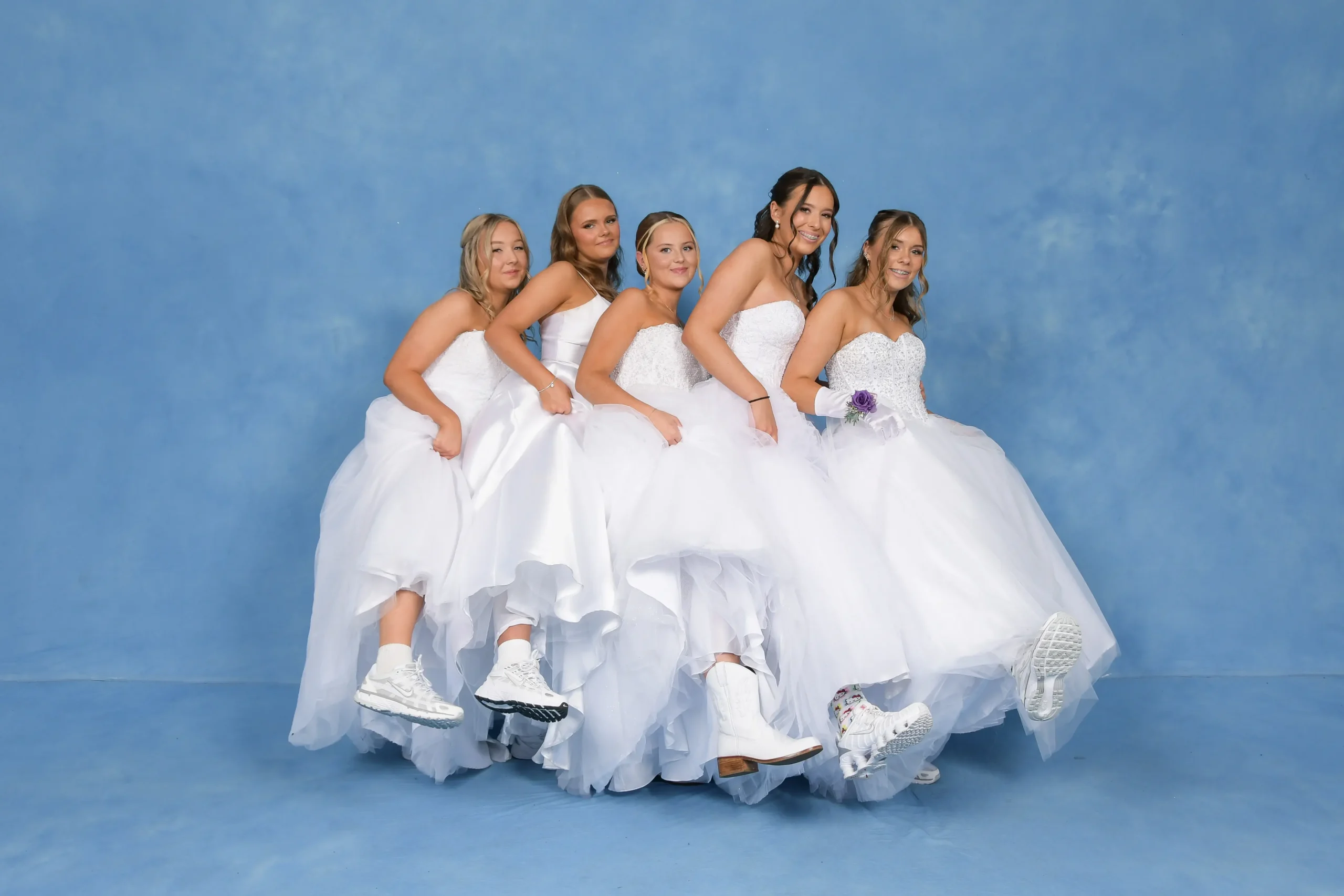 Five debutantes in white dresses against a blue backdrop, lifting their skirts to reveal casual white sneakers and boots.