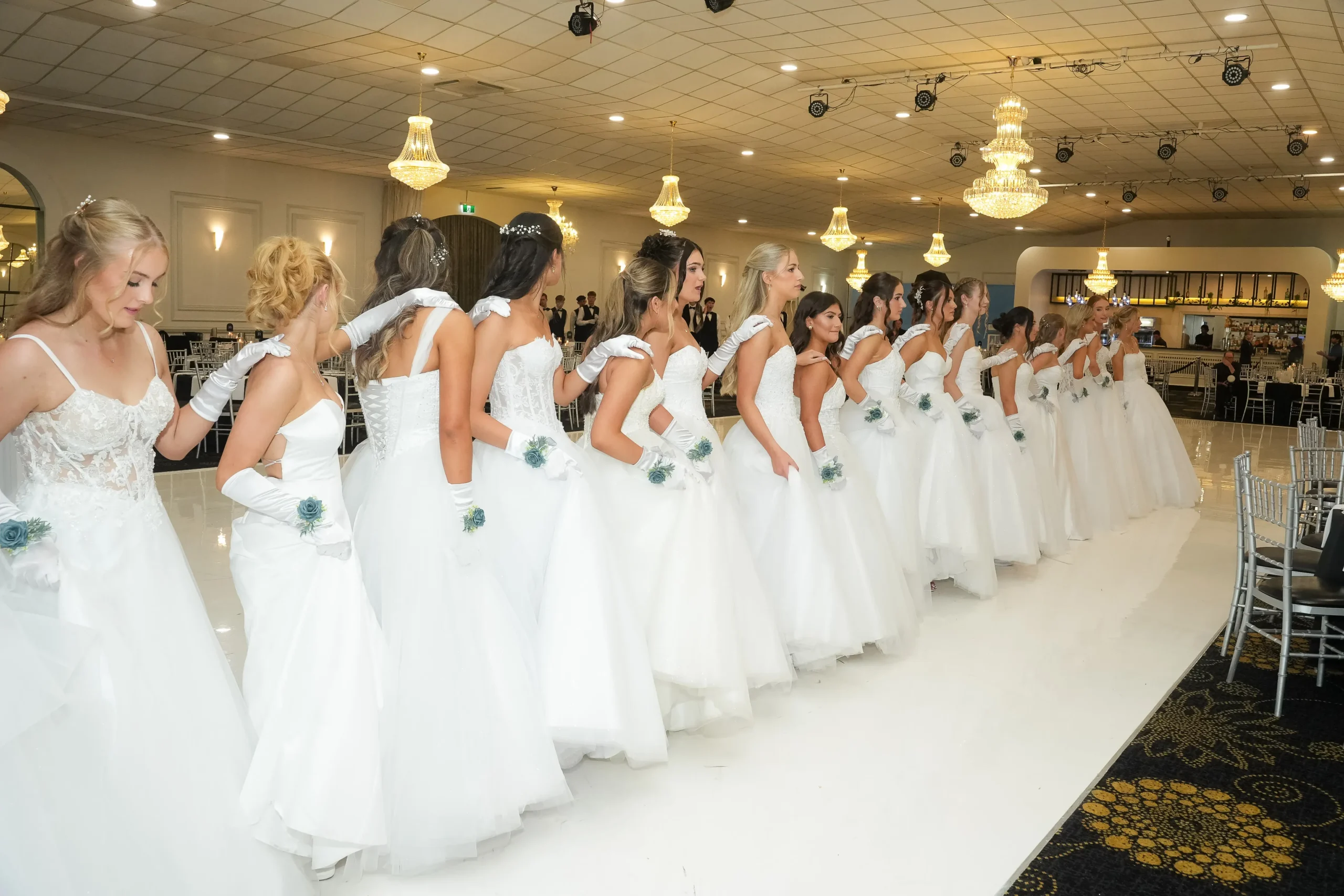 A long line of debutantes in white dresses standing in a conga line formation on a white ballroom dance floor.