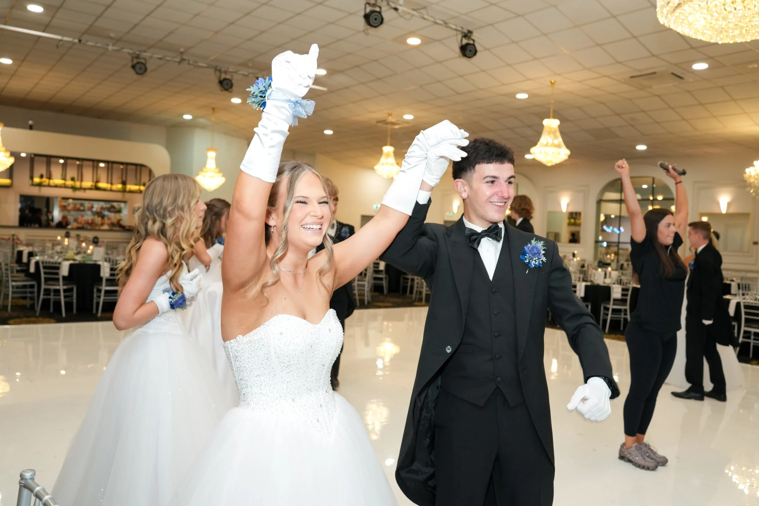 A smiling couple in formal debutante attire raising their joined hands in celebration on a brightly lit dance floor.