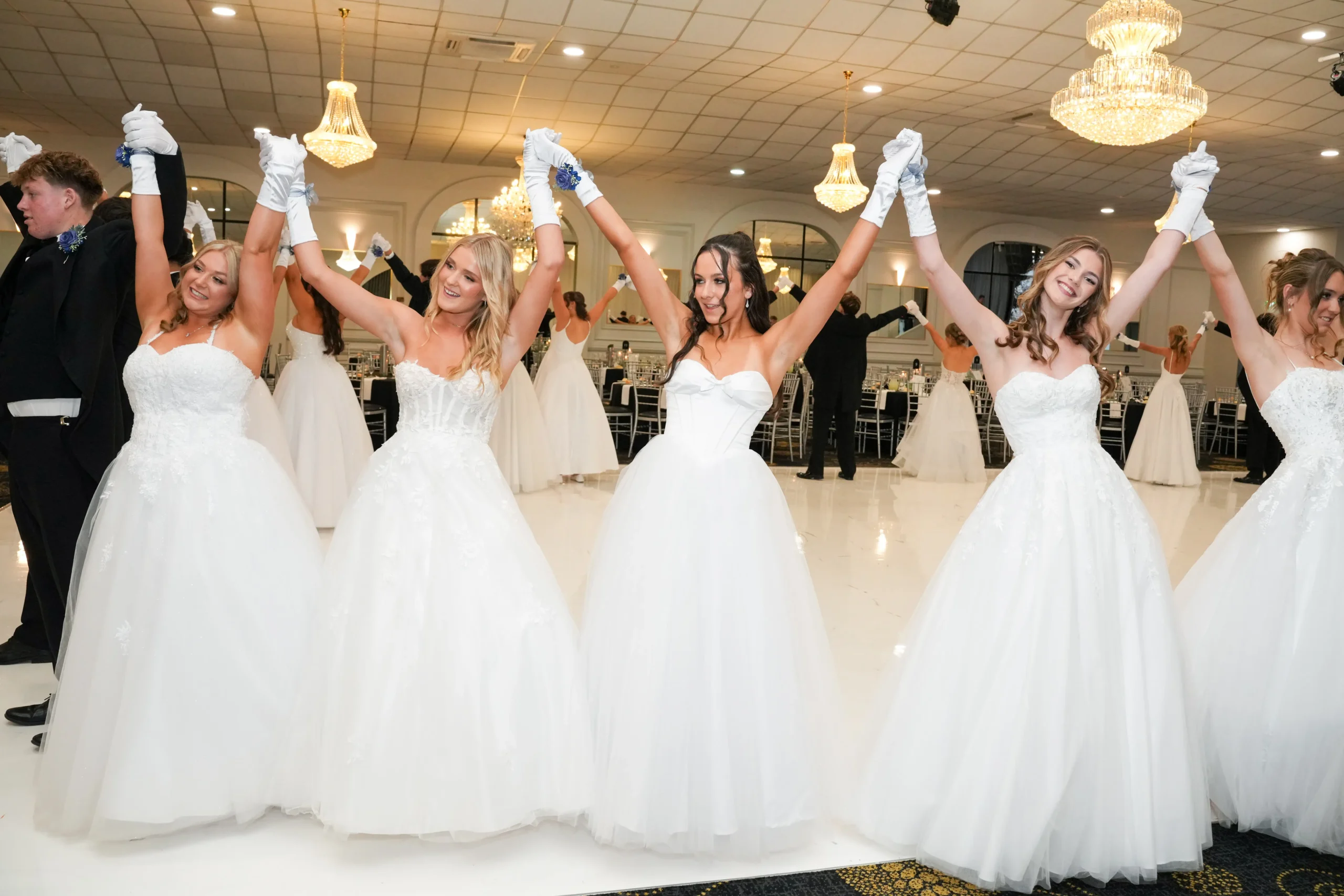 A line of debutantes in white gowns and their partners in black suits raising their arms together on a brightly lit dance floor.