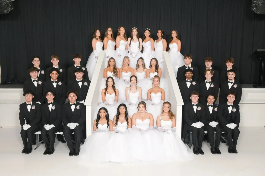A large formal group portrait of debutantes in white gowns and their partners in black suits posed on white steps.