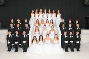 A large formal group portrait of debutantes in white gowns and their partners in black suits posed on white steps.