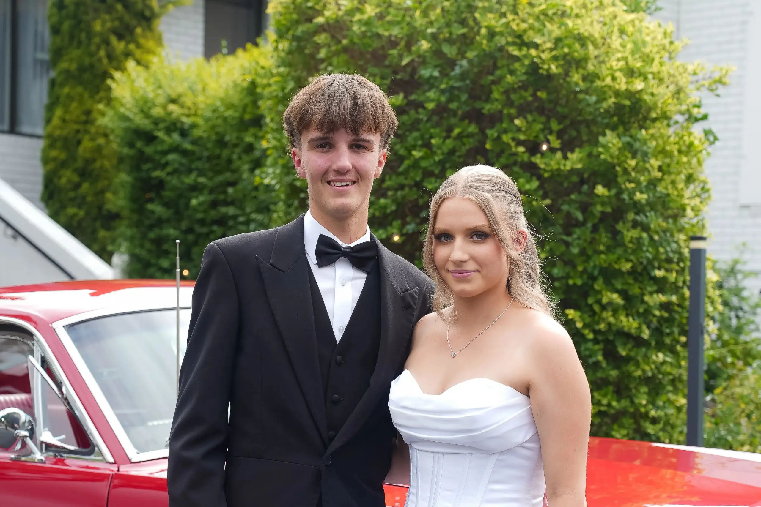 Debutante and partner in formal attire posing outdoors beside a red car.