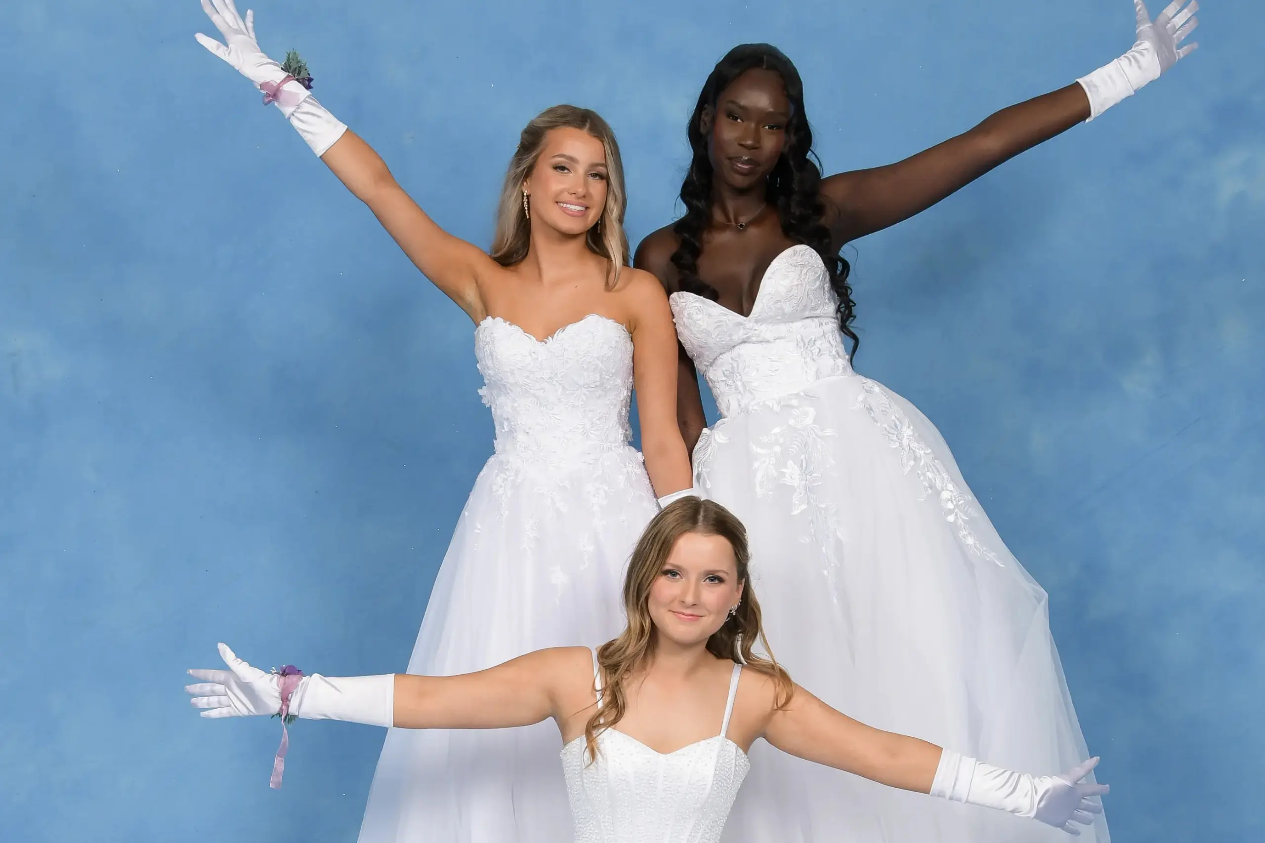 Three debutantes in white gowns posing playfully with extended arms against a blue background.